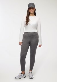 Top a maniche lunghe bianco, leggings grigi elasticizzati e un cappellino grigio con accenti brillanti. Modella in scarpe sportive su uno sfondo neutro.