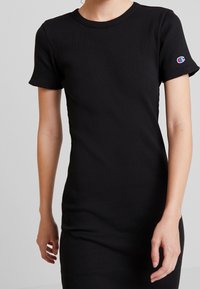 Robe t-shirt noire côtelée avec des manches courtes, un col rond et un petit logo brodé sur la manche gauche. Texture lisse.