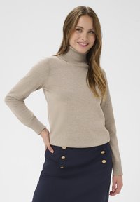 Pull beige à col roulé avec manches longues et poignets côtelés, associé à une jupe bleu marine ornée de boutons dorés sur le côté.