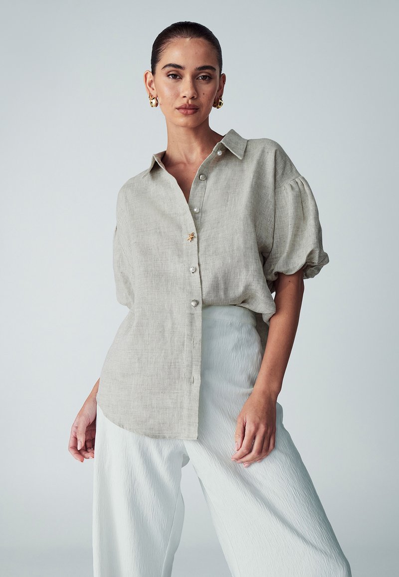Light beige linen shirt with short puff sleeves, front button closure, and a soft texture, paired with white pleated wide-leg trousers.