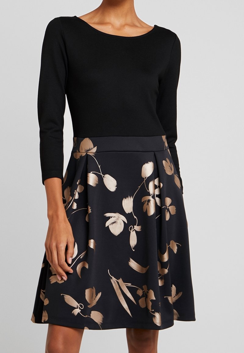 Femme portant une robe noire à manches trois-quarts avec une jupe beige à motif floral, debout devant un fond uni gris clair.