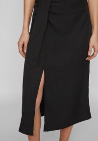 Jupe noire mi-longue avec une fente sur le côté, en tissu texturé. Le design présente un nœud drapé à la taille et des bords lisses.