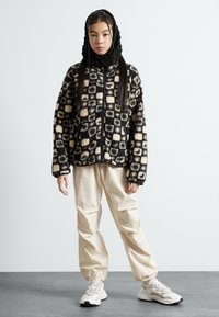 Zwart en bruin geruite fleecejas met rits aan de voorkant, gecombineerd met losse crème broek en witte sneakers. Effen zwarte capuchon zichtbaar.