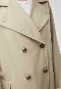 Beige double-breasted coat with large, shiny buttons, wide lapels, and a smooth fabric texture. Features detailed stitching and pockets.