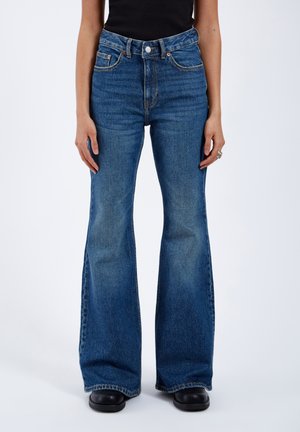 Person trägt hoch taillierte, mittelblaue Schlagjeans, schwarzen Oberteil und schwarze Schuhe, steht vor einem schlichten hellen Hintergrund.