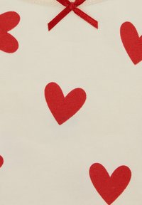 Bombažno blago s vzorcem rdečih src na krem ozadju in rdečo pentljo ob izrezu vratu.