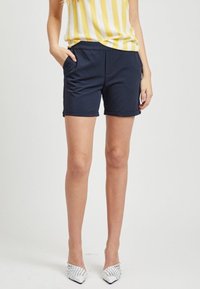 Shorts bleu marine en tissu extensible, dotés d'une taille élastique et de poches latérales. Portés avec un haut à rayures jaunes et des chaussures blanches.