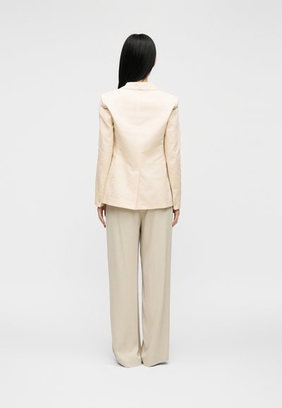 Femme aux longs cheveux noirs debout, portant un blazer beige et un pantalon à jambes larges, vue de dos sur un fond clair uni.