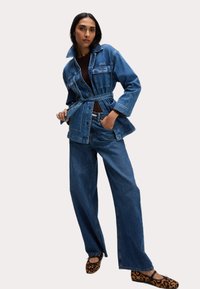 Giacca di jeans con vita regolabile, dotata di tasche frontali, colletto e chiusure a bottoni, abbinata a pantaloni di jeans a gamba larga e scarpe con stampa animale.