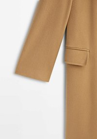 Manche et poche à rabat d'un manteau en laine couleur camel avec une texture lisse sur fond blanc.