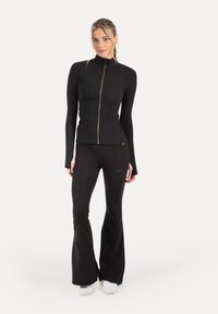 Chaqueta atlética negra con cuello alto y cremallera dorada, combinada con pantalones negros acampanados. Textura suave, diseño ajustado y marca sutil.