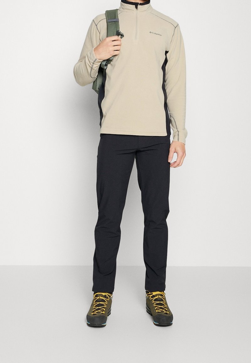 Homme portant un pull beige Columbia, un pantalon noir, des chaussures de randonnée vertes avec des lacets jaunes, et portant une sangle de sac à dos verte sur une épaule.
