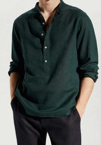 Uomo che indossa una maglietta henley a maniche lunghe verde scuro con tre bottoni, con le maniche arrotolate, mani nelle tasche, in piedi contro uno sfondo neutro.
