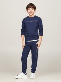 Dunkelblaues Sweatshirt mit dem Logo "TOMMY HILFIGER EST. 1985", kombiniert mit passenden Jogginghosen. Weiße Sneakers mit navyfarbenen Akzenten vervollständigen das Outfit.