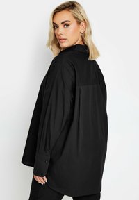 Camisa negra de gran tamaño con cuello, mangas largas y puños con botones. Presenta un detalle de canesú en la parte posterior y una tela suave y ligera.