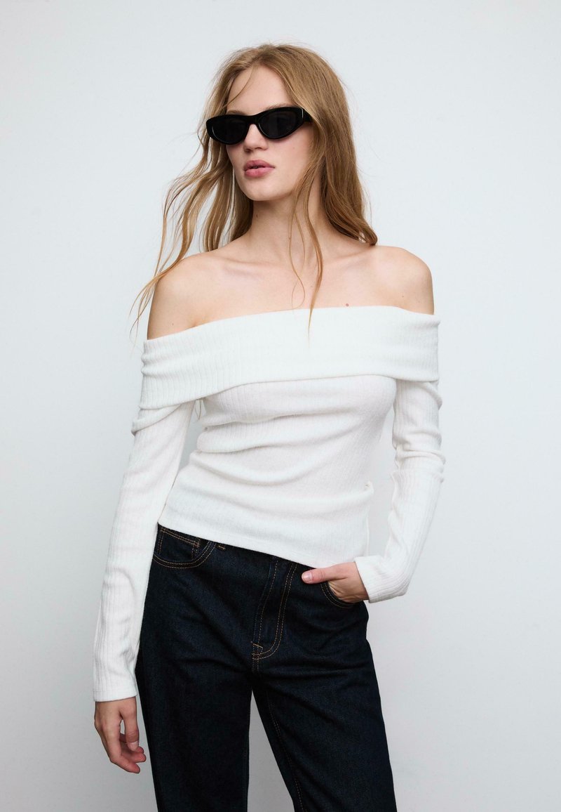 Bílý off-shoulder ribboný top s dlouhými rukávy, doplněný tmavými denimovými kalhotami s viditelným šitím. Modelka nosí černé sluneční brýle.