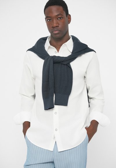 Witte button-up shirt en lichtblauwe gestreepte broek. Donker breiwerk vest om de schouders geknoopt. Glad stoftexturen en strakke lijnen.