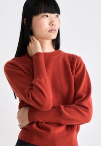 Junge Frau mit langen schwarzen Haaren, die einen roten Strickpullover trägt, hält sich mit einer Hand den Hals und hat den anderen Arm über ihre Taille gekreuzt.