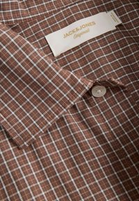 Καφέ πουκάμισο καρό με μαλακό ύφασμα, διαθέτοντας λευκές γραμμές πλέγματος, κόψη αιχμής και μια λεπτομέρεια με ένα κουμπί στην πλάκα.