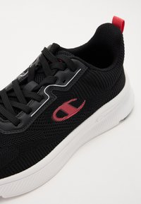 Scarpa sportiva nera in tessuto a rete, con accenti rossi e suola bianca. Presenta lacci e logo sul lato. Dettaglio della superficie testurizzata.