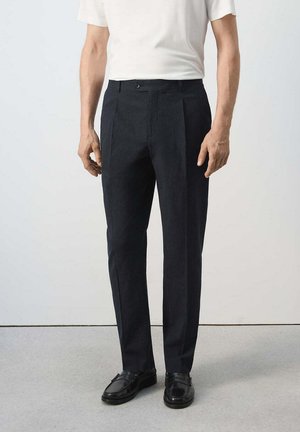 Uomo che indossa pantaloni eleganti neri e mocassini neri in piedi contro un muro bianco semplice e pavimento grigio.