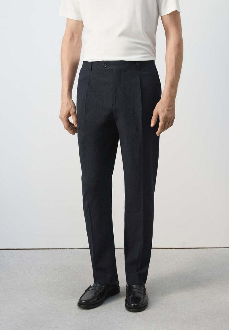 Homme portant un pantalon de costume noir et des mocassins noirs, debout contre un mur blanc simple et un sol gris.