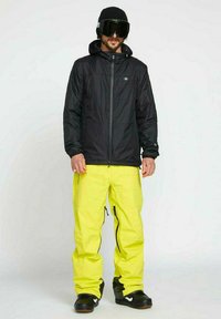 Schwarze isolierte Jacke, gelbe Schneehose, schwarze Stiefel. Die Jacke verfügt über eine Kapuze und einen Reißverschluss vorne; die Hose hat Reißverschlüsse an den Seiten.