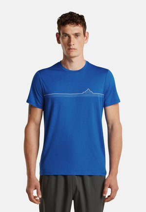 Mann trägt ein blaues T-Shirt mit kurzen Ärmeln mit einer weißen horizontalen Linie und Bergmotiv auf der Brust, steht vor einem schlichten Hintergrund.