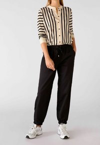 Pruhovaná košile na knoflíky v krémové a černé barvě, spárovaná s černými kalhotami s elastickým pasem. Model nosí bílé sportovní tenisky.