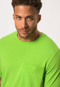 Světle zelené tričko s krátkým rukávem, kulatým výstřihem a náprsní kapsou na levé straně. Hladká textura, neformální design.