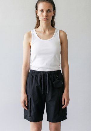 Young woman with wet hair wearing a white sleeveless top and black drawstring cargo shorts with a small pouch attached.