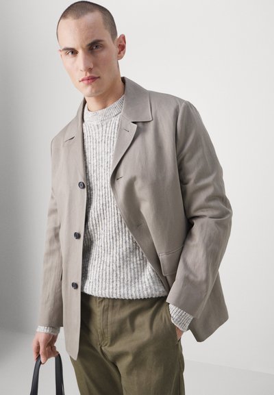 Lichtgrijze blazer met een gestructureerd ontwerp, gecombineerd met een textuur crème trui en olijfgroene broek, vastgehouden met een zwarte tas.