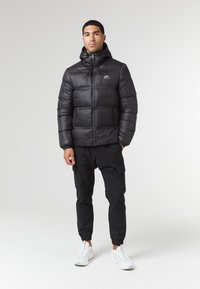 Giubbotto imbottito nero con cappuccio, dotato di un design a quadri e tasche con zip. Abbinato a pantaloni cargo neri e sneaker bianche.