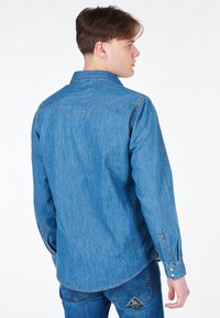 Camicia in denim di colore azzurro chiaro con colletto, maniche lunghe e polsini con bottone. Presenta uno sprone triangolare e una piega sul retro per una vestibilità comoda.