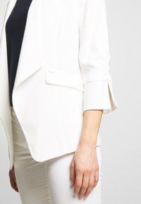 Blazer bianco con frontale drappeggiato, maniche a tre quarti e una sottile fessura al polsino. Abbinato a una maglia blu navy e pantaloni bianchi.