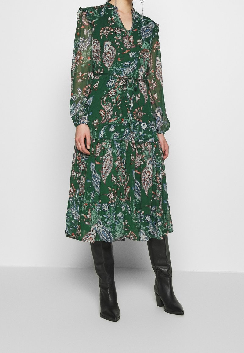 Mujer vestida con un vestido midi de manga larga de estampado paisley verde, con un lazo en la cintura y botas de tacón negras hasta la rodilla.