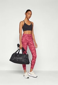 Juodas sportinis liemenėlė, raudonos gėlių rašto tamprės ir balti sportbačiai. Vienoje rankoje laikoma juoda kelioninė kuprinė. Rodoma prigludusi atletinė stiliaus apranga.