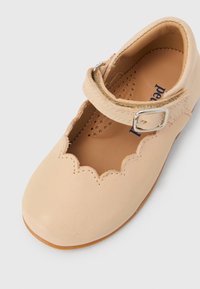 Μπεζ δερμάτινο παπούτσι Mary Jane με κυματιστό περίγραμμα, διατρητό πέλμα και ρυθμιζόμενο λουράκι με ασημένια αγκράφα σε λευκό φόντο.