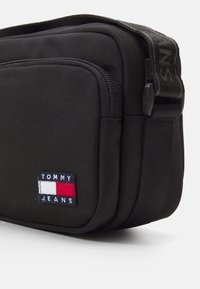 Borsa a tracolla in tessuto nero con tasca frontale con zip, caratterizzata da un patch con il logo Tommy Jeans in blu, bianco e rosso. Superficie texture, design compatto.