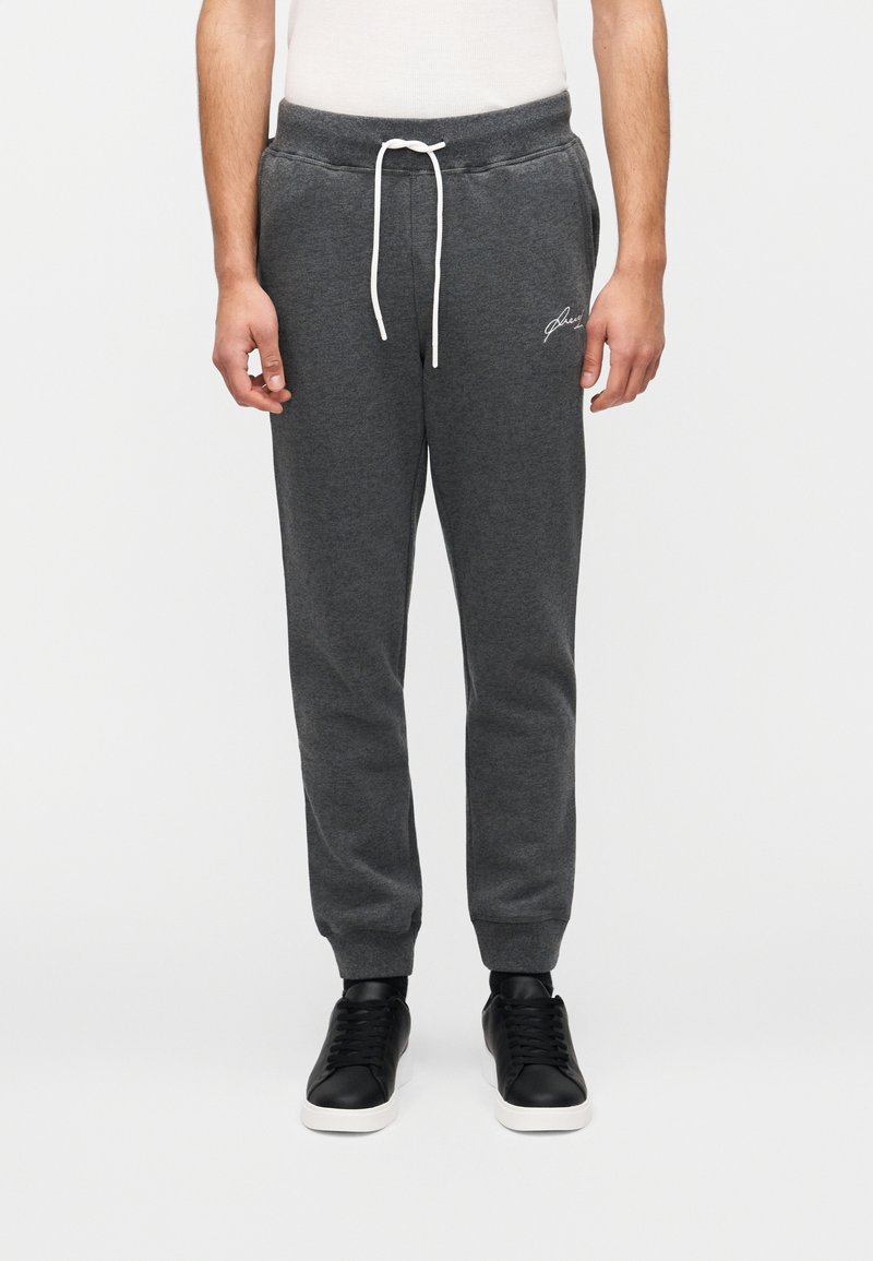 Γκρι φόρμα με κορδόνι στη μέση, πλαϊνές τσέπες και κεντημένο λογότυπο στον αριστερό μηρό, σε συνδυασμό με μαύρα αθλητικά παπούτσια.