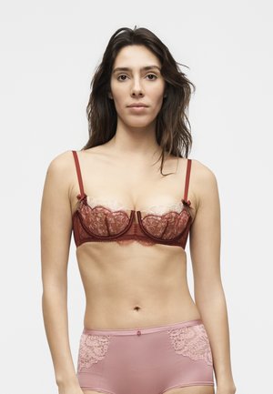 Mujer con cabello largo y oscuro, vistiendo un sujetador de encaje burdeos y ropa interior de talle alto rosa con ribete de encaje, de pie frente a un fondo blanco liso.