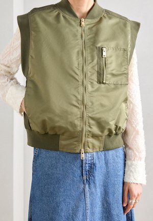 Persona con un chaleco sin mangas verde oliva con cremallera y bolsillo en el pecho sobre una blusa blanca de manga larga texturizada y una falda vaquera azul.