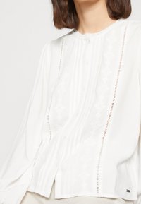 Blusa bianca a maniche lunghe con chiusura a bottoni, design plissettato, accenti ricamati e dettagli a intaglio delicati lungo le cuciture.