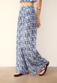 Vrouw wandelt in een wijde blauwe en witte gebloemde broek, zwarte sandaal en een gouden armband om haar arm tegen een effen achtergrond.