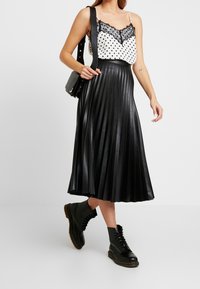 Gonna midi plissettata nera con cintura aderente, abbinata a una blusa bianca con pois neri e rifiniture in pizzo nero. Indossata con stivali alla caviglia neri con lacci.