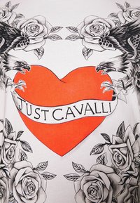 Röd hjärta med en "Just Cavalli"-banner, omgiven av svarta och vita rosor samt två örnar som står mitt emot varandra med öppna näbbar och uppsträckta klor.