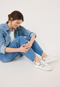 Sneakers bianchi con accenti grigi, realizzati in un materiale liscio, con punta tonda e suola texturizzata. Indossati con jeans blu chiaro e giacca di jeans.