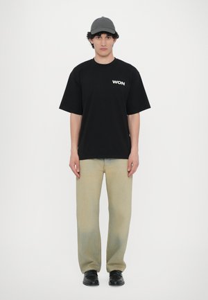 Svart bomulls-t-shirt med "WON" tryckt i vitt, kombinerad med ljusbeige denimbyxor och en grå keps. Modellen står framför en enfärgad bakgrund.