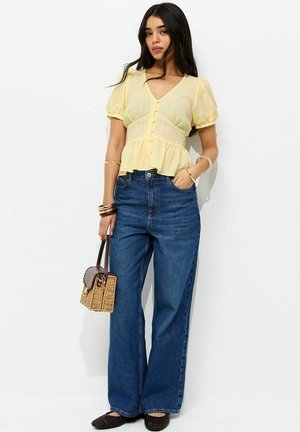 Femme portant un chemisier jaune clair à manches bouffantes, un jean bleu taille haute à jambes larges, tenant un petit sac tressé, avec des bracelets aux poignets.