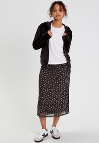 Haut à zip noir sur un t-shirt blanc, associé à une jupe midi noire à imprimé floral et des sneakers blanches avec des accents foncés.